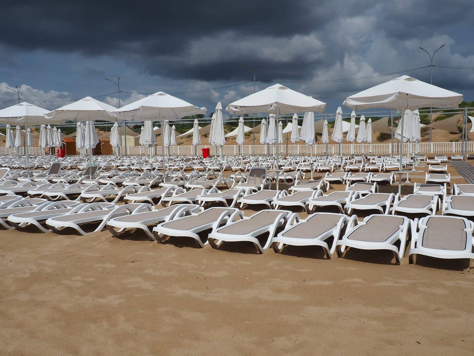 Wet sunbeds and umbrellas on the sea beach during heavy rain storm. Rolled up parasol on the beach against dark cloudy sky. Invasion of a typhoon, cyclone or storm on the sea or ocean. Beach season
