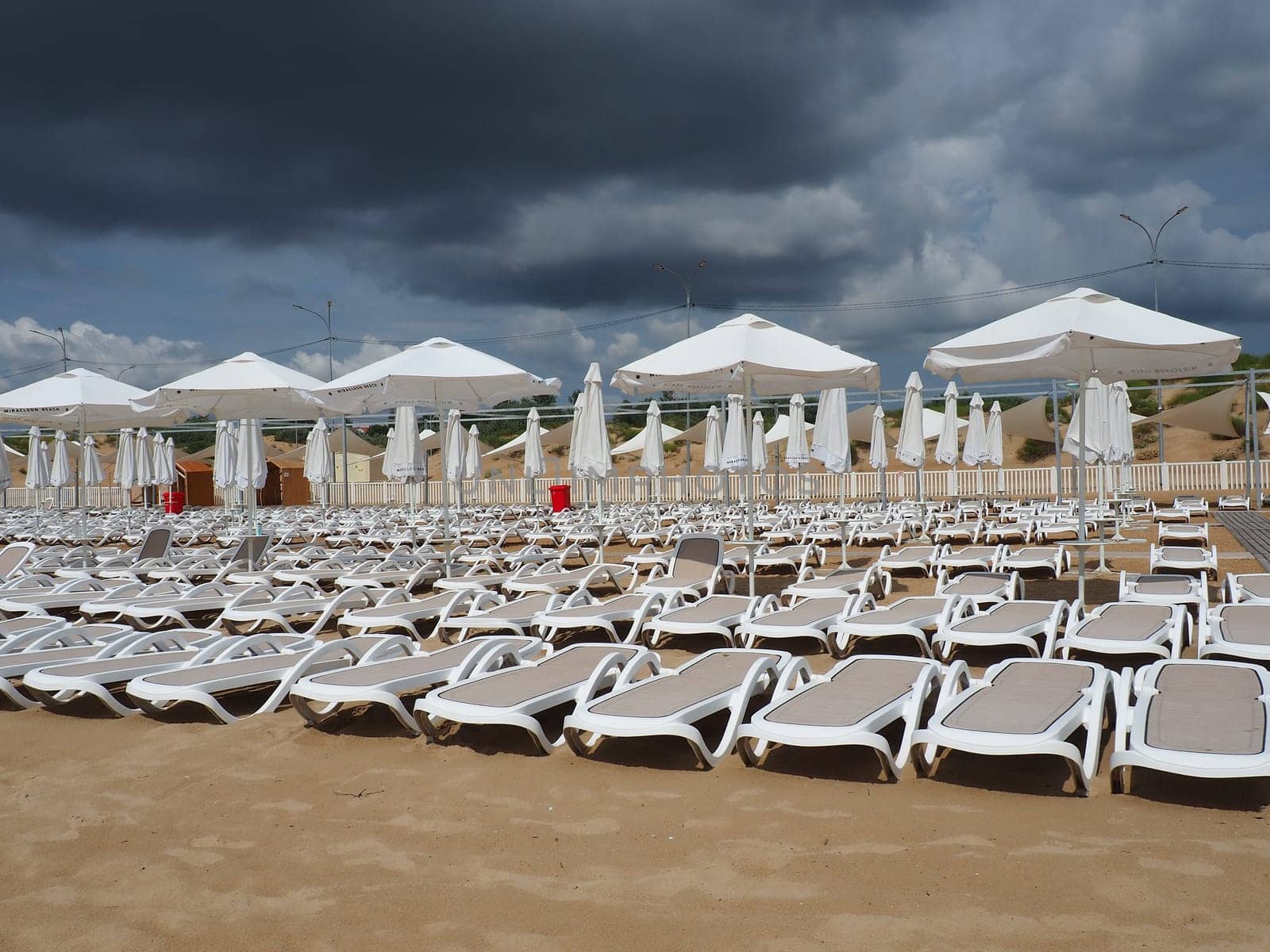 Wet sunbeds and umbrellas on the sea beach during heavy rain storm. Rolled up parasol on the beach against dark cloudy sky. Invasion of a typhoon, cyclone or storm on the sea or ocean. Beach season