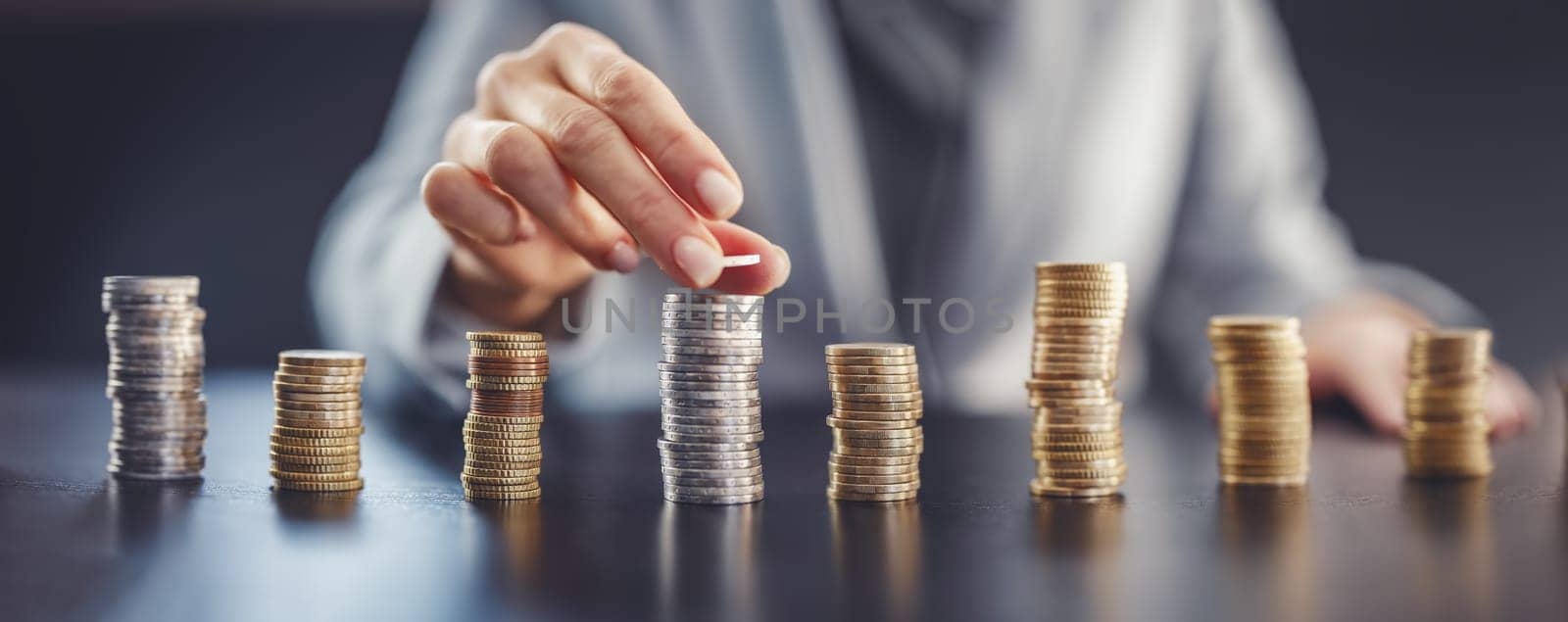 Woman sitting at the table and putting coin on the largest stack of the money. Concept of the wages and financial income in business.