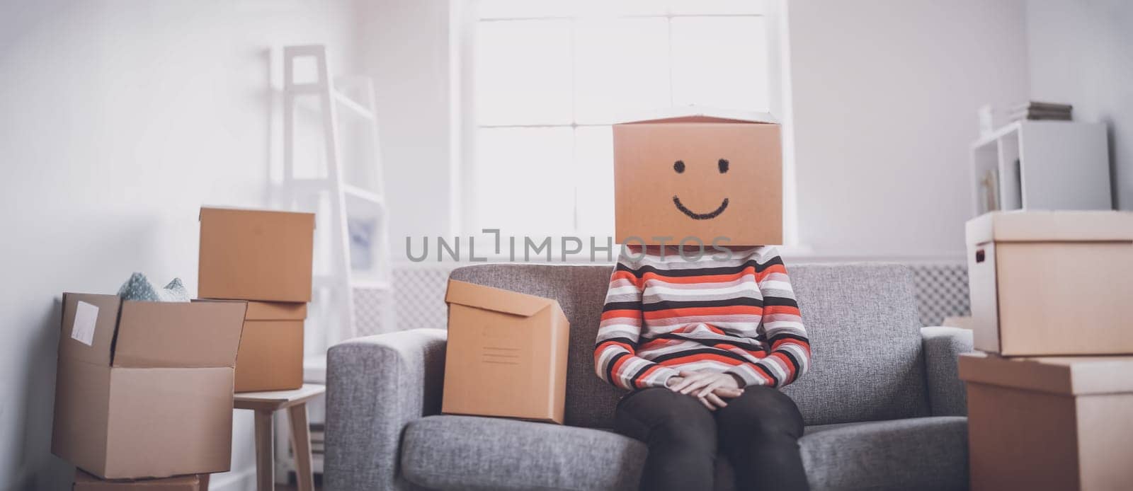 Tired happy woman with cardboard box on her head sitting on the sofa in her new house. Concept of renting, moving in new house and buying an real estate.
