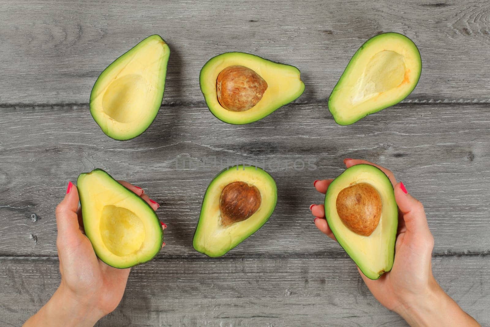 Tabletop view, woman hands holding avocado cut in half, seed visible, more avocados halves on gray wood desk around.