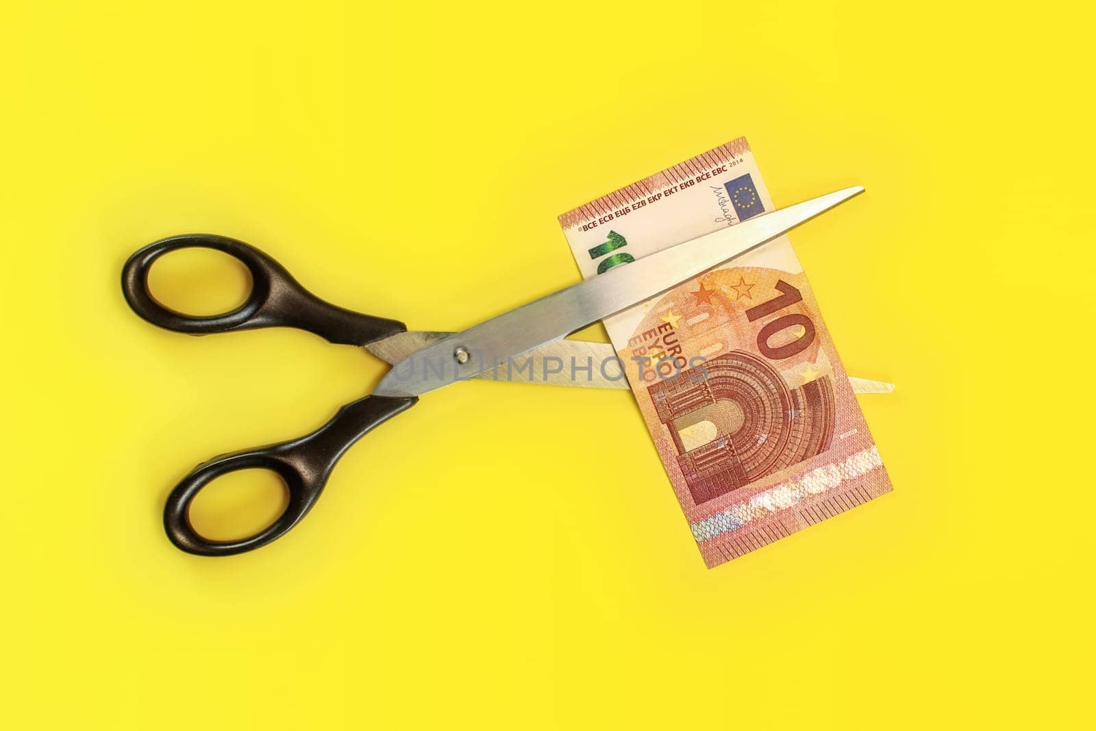 Tabletop view - large scissors cutting ten eur banknote over yellow board. Cut fees concept.