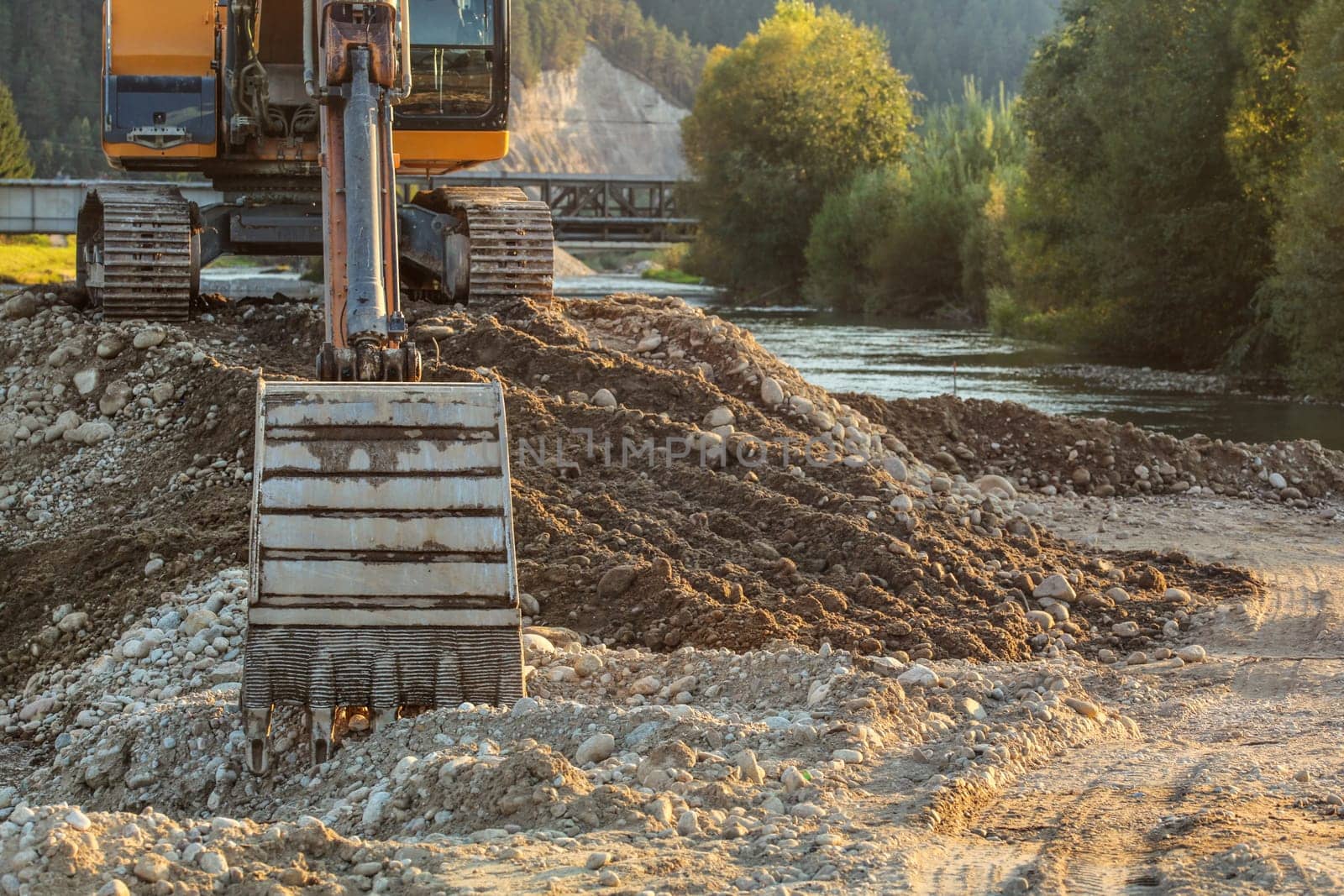 Small yellow excavator at pile of rocks and stones next to river, detail on digger bucket on ground. Construction at riverside.