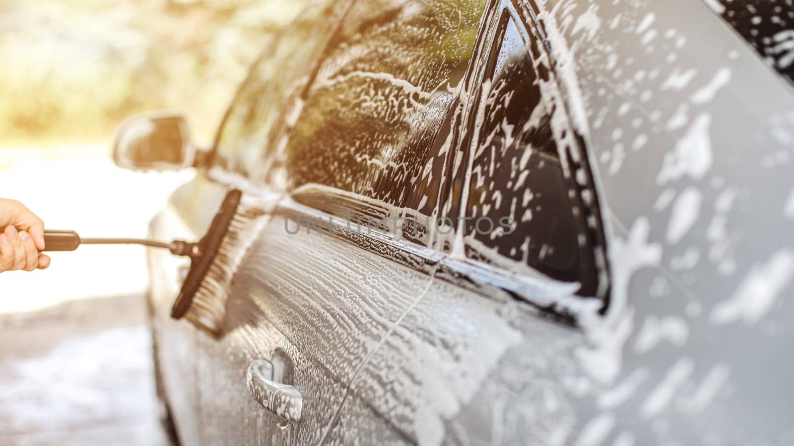 Side of silver car washed in self serve carwash, brush held by man's hand leaving strokes in shampoo, sun backlight in background.
