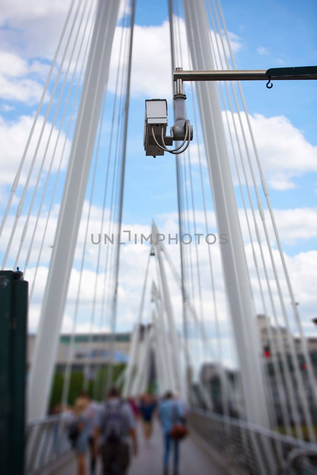 Detail of surveillance camera with unrecognisable people walking by in blurred background, and blue sky above.