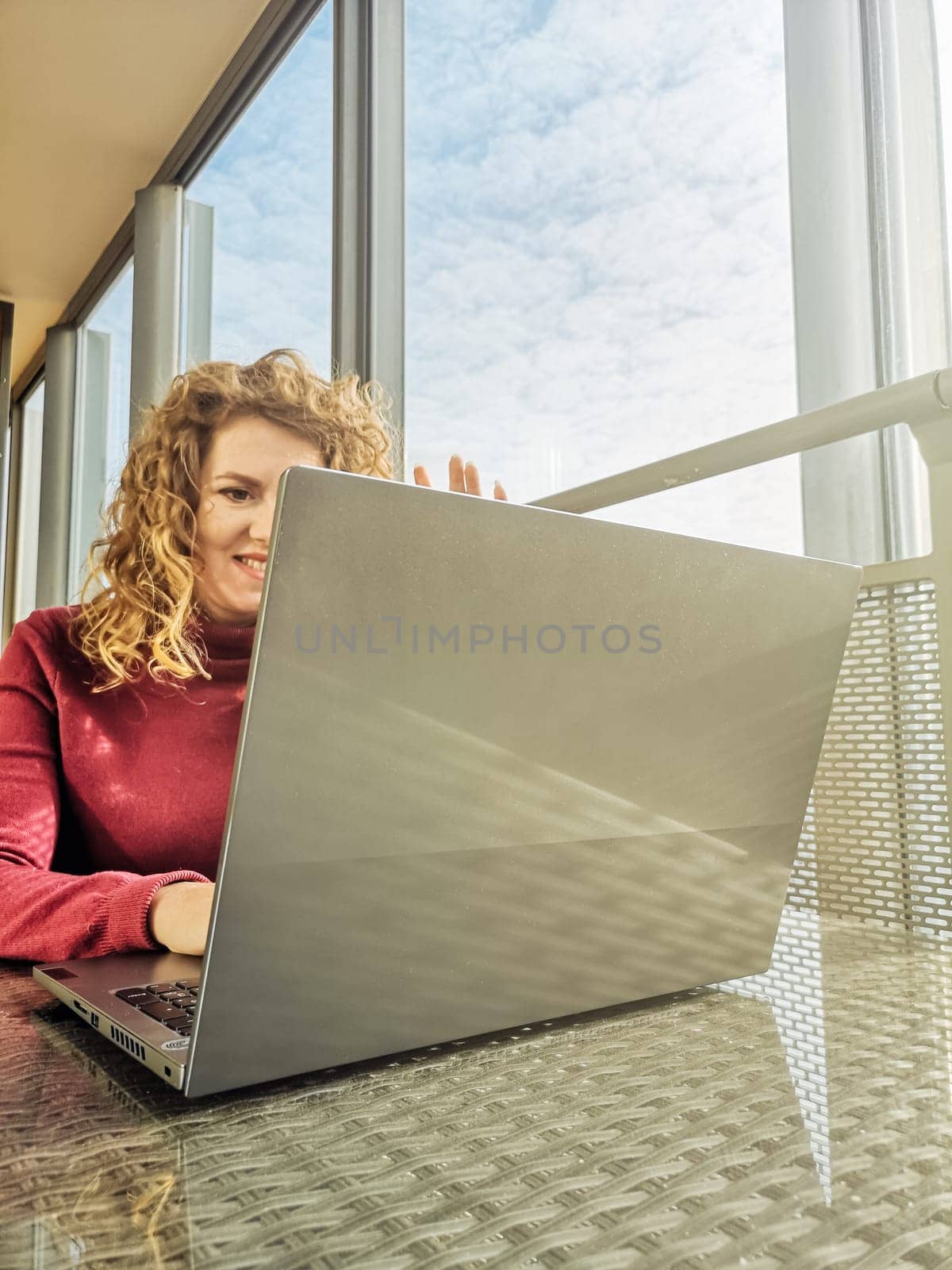 A blonde with curly hair communicates via video link on a laptop sitting in front of a window.