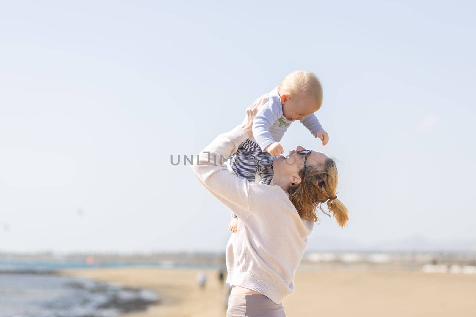 Mother enjoying summer vacations holding, playing and lifting his infant baby boy son high in the air on sandy beach on Lanzarote island, Spain. Family travel and vacations concept