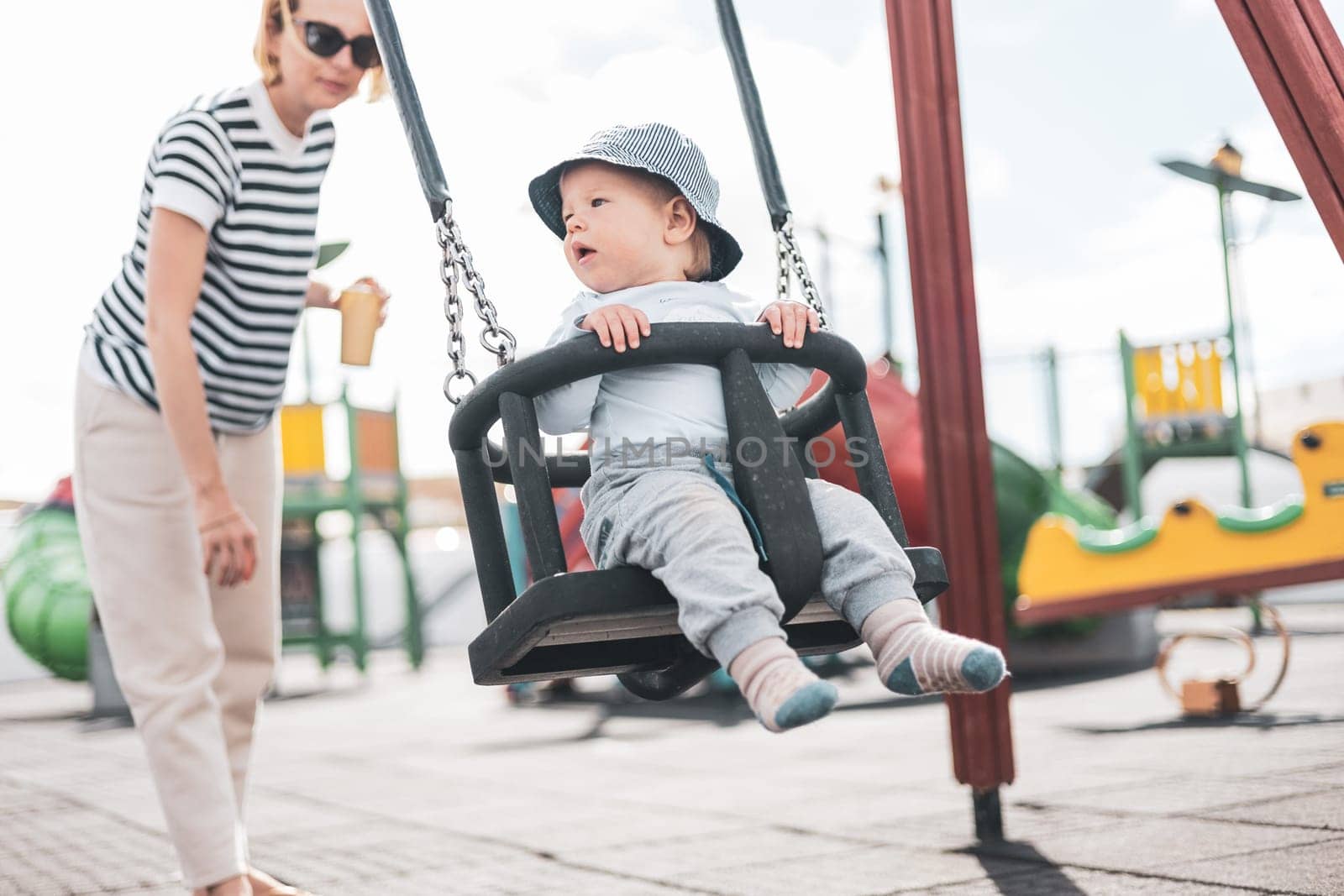 Mother pushing her infant baby boy child on a swing on playground outdoors.