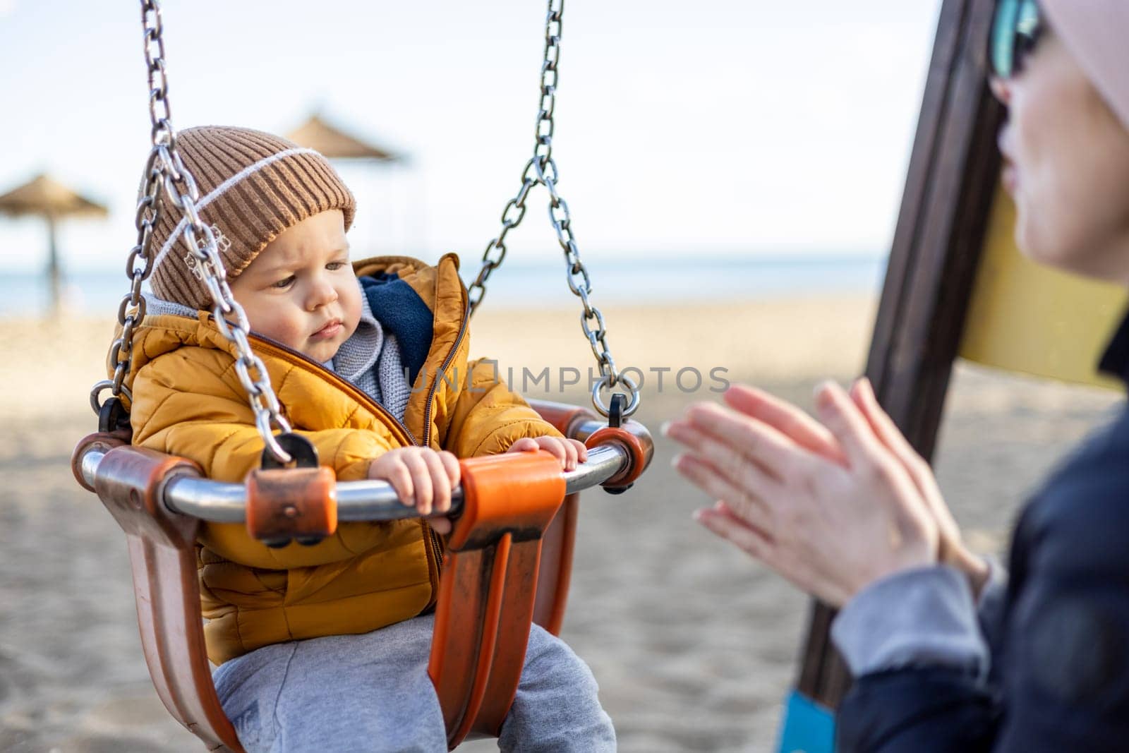 Mother pushing her infant baby boy child on a swing on sandy beach playground outdoors on nice sunny cold winter day in Malaga, Spain