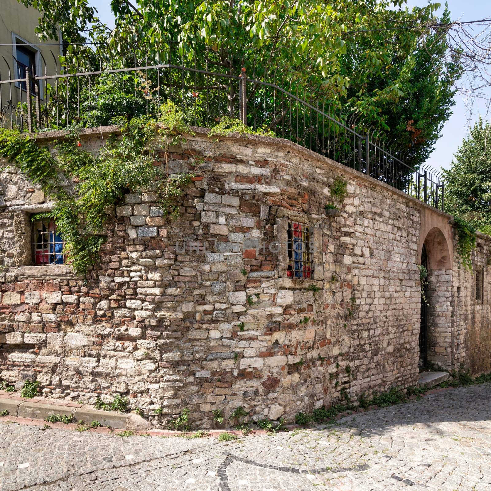 Old abandoned building, with weathered stone wall, and wrought iron windows, partially covered with climber plants, in a narrow alley with cobblestone floor, Balat district, Istanbul, Turkey