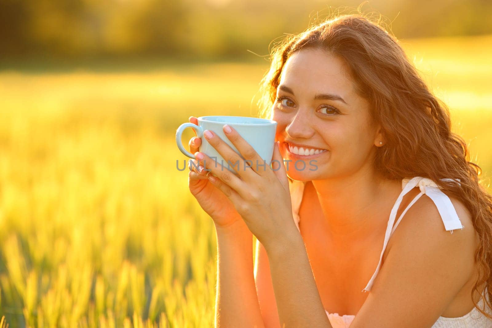 Happy woman in a field looking at you drinking coffee at sunset