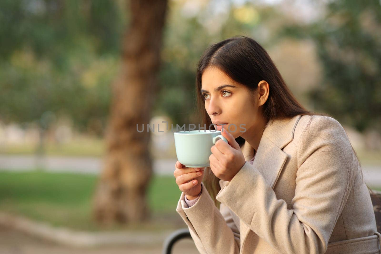 Woman drinking coffee looks away at sunset in a park in winter
