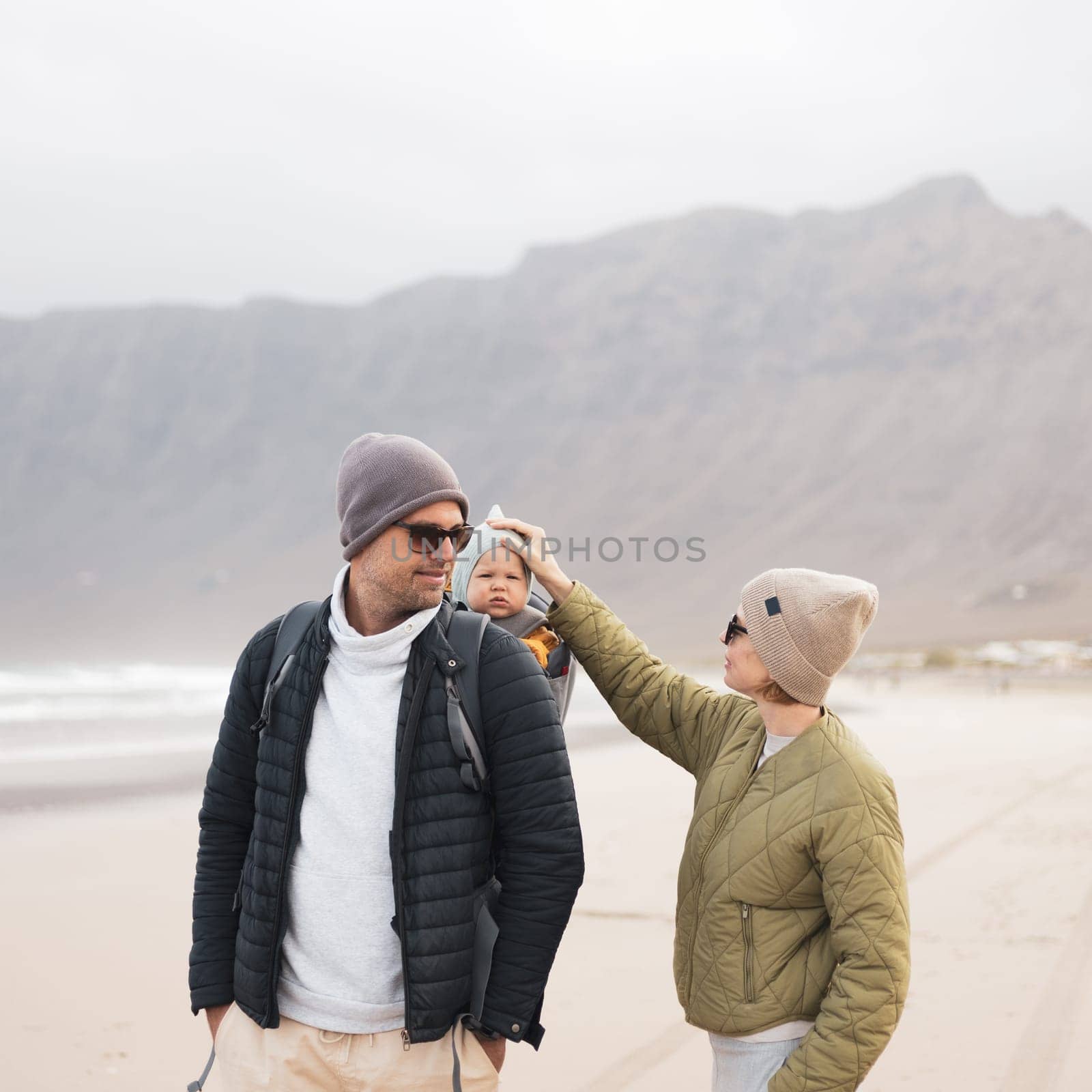 Young father carrying his infant baby boy son in backpack on windy sandy Famara beach, Lanzarote island, Spain. Family travel and winter vacation concept