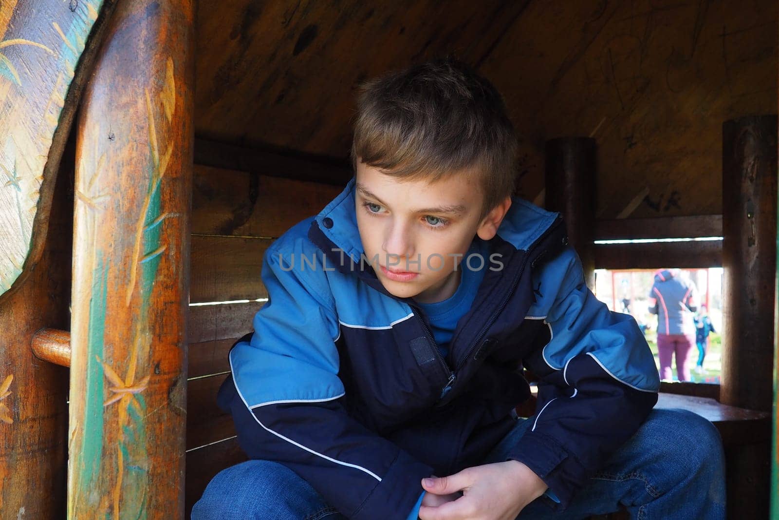 A 10-year-old Caucasian boy with blond hair and gray eyes looks out of the door of a wooden house on a children's playground. Peekaboo. The child hid. Nice face. The boy is sitting in a small house