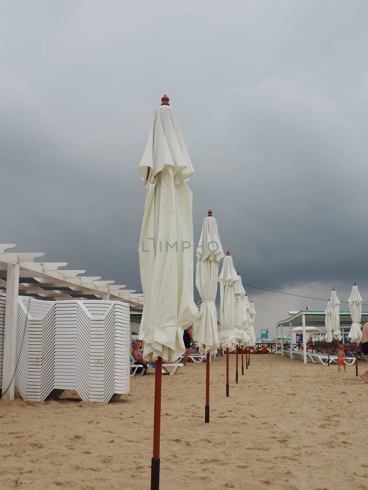 Wet sunbeds and umbrellas on the sea beach during heavy rain storm. Rolled up parasol on the beach against dark cloudy sky. Invasion of a typhoon, cyclone or storm on the sea or ocean. Beach season.