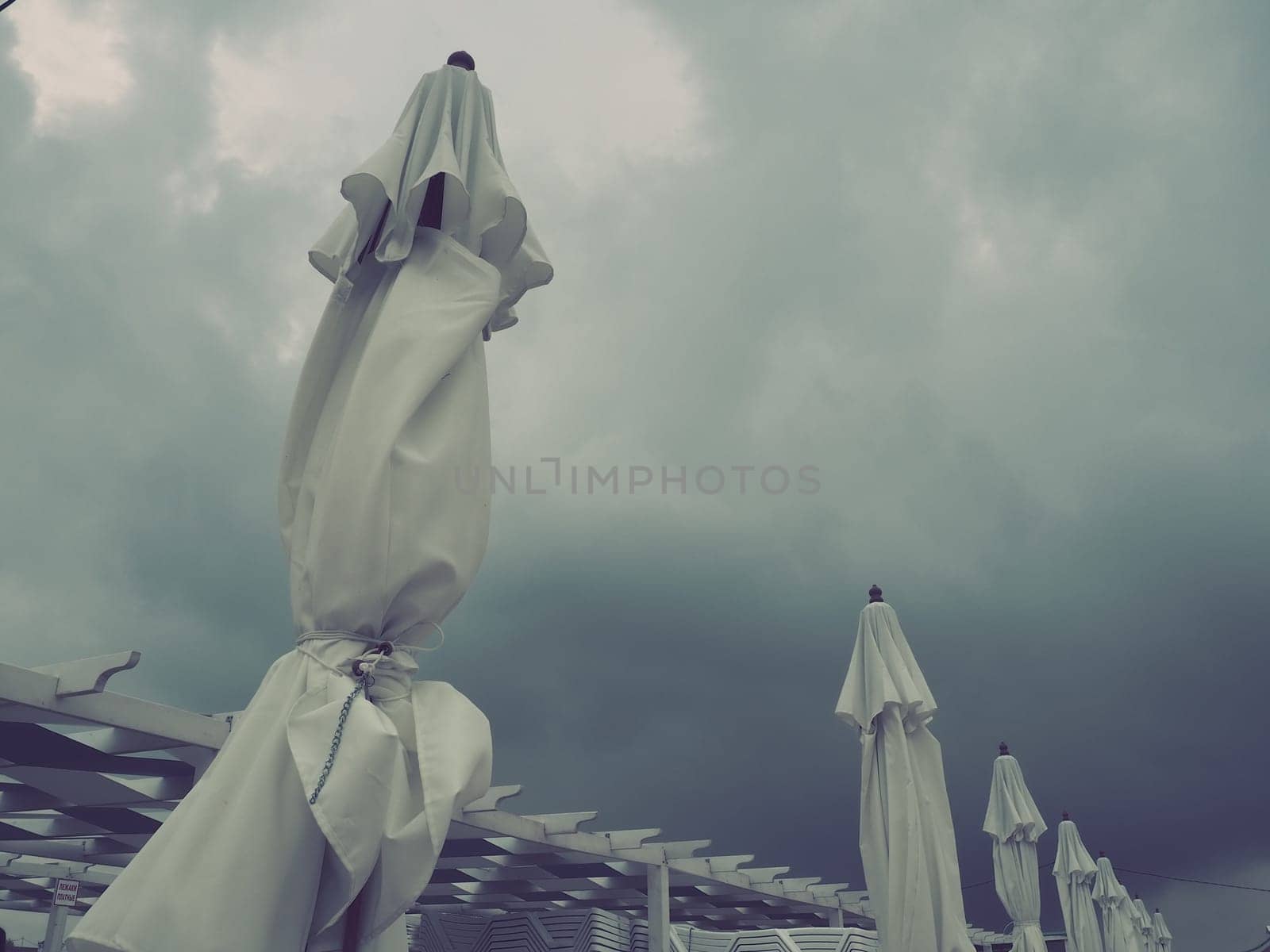 Wet sunbeds and umbrellas on the sea beach during heavy rain storm. Rolled up parasol on the beach against dark cloudy sky. Invasion of a typhoon, cyclone or storm on the sea or ocean.