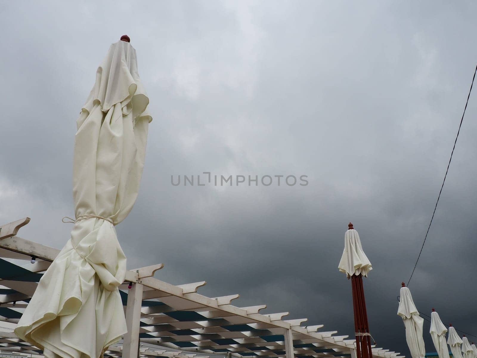 Wet sunbeds and umbrellas on the sea beach during heavy rain storm. Rolled up parasol on the beach against dark cloudy sky. Invasion of a typhoon, cyclone or storm on the sea or ocean. Beach season.