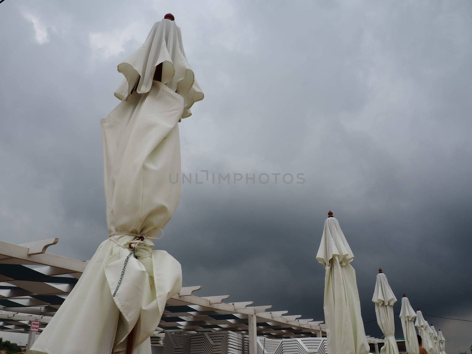 Wet sunbeds and umbrellas on the sea beach during heavy rain storm. Rolled up parasol on the beach against dark cloudy sky. Invasion of a typhoon, cyclone or storm on the sea or ocean. Beach season.