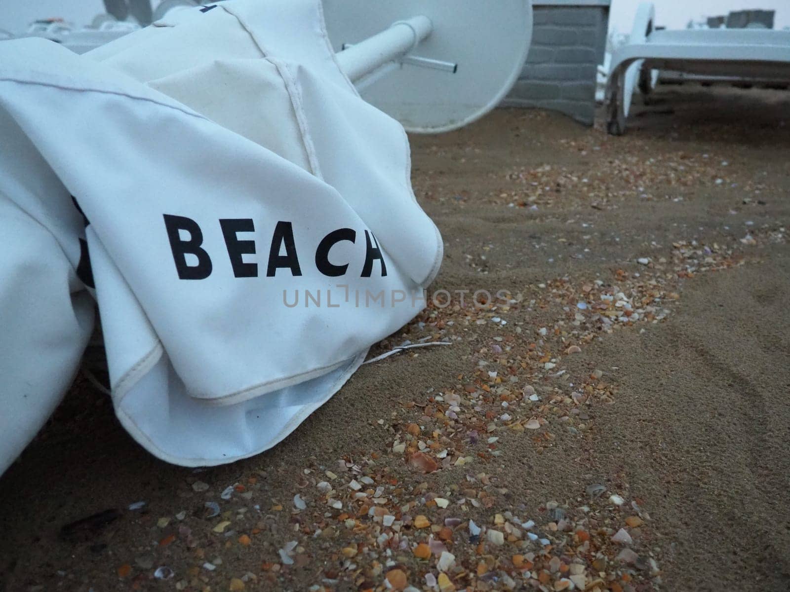 A beach umbrella lay folded on a sandy beach after bad weather. Storm at sea. End of the beach season due to typhoon and cyclone. Loungers, umbrellas and hotel furniture. Beach word written on white.