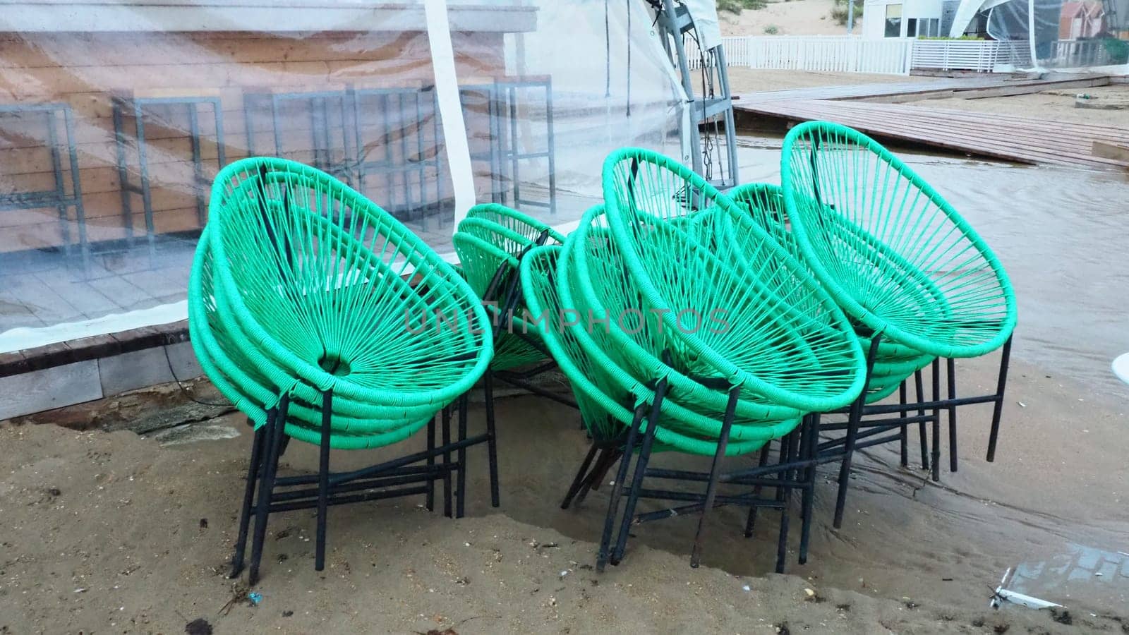 wicker green plastic chairs with black legs in a cafe on a sandy beach after a storm. The furniture is stacked on top of each other as the beach cafe is closed. End of the beach season at the hotel.