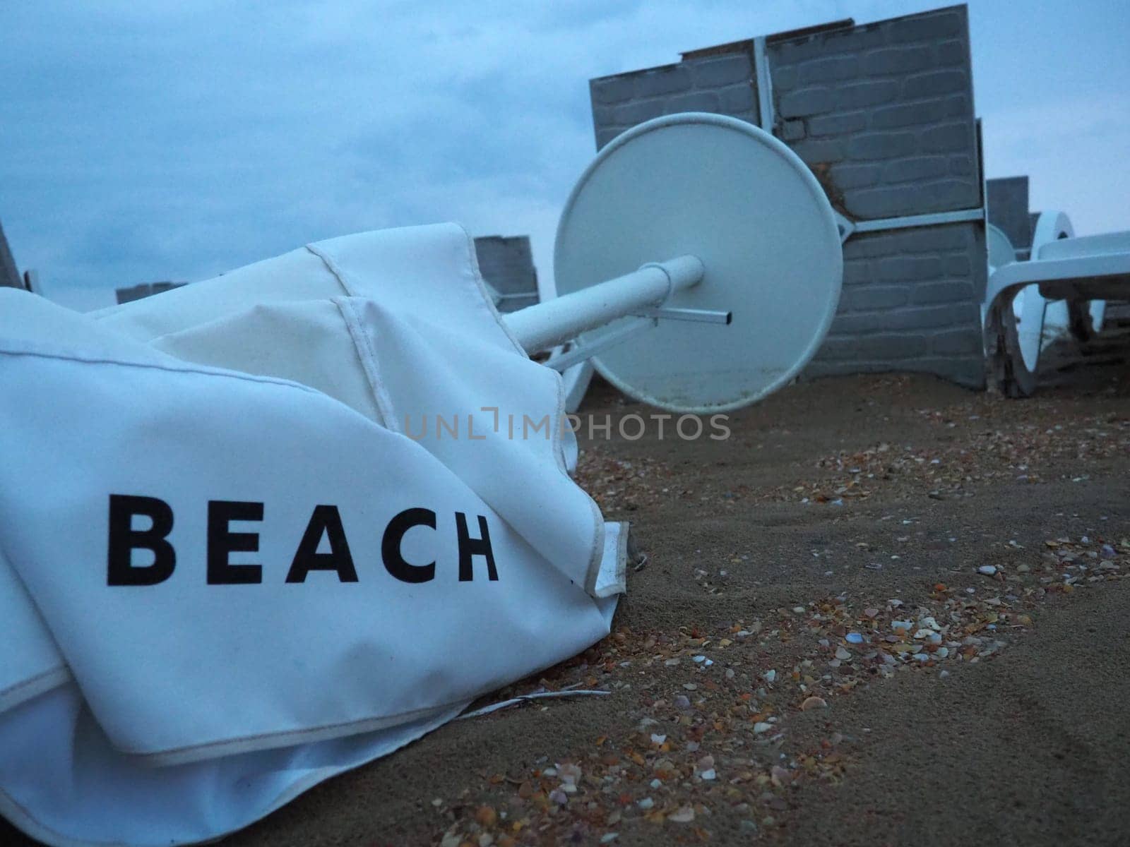 A beach umbrella lay folded on a sandy beach after bad weather. Storm at sea. End of the beach season due to typhoon and cyclone. Loungers, umbrellas and hotel furniture. Beach word written on white.