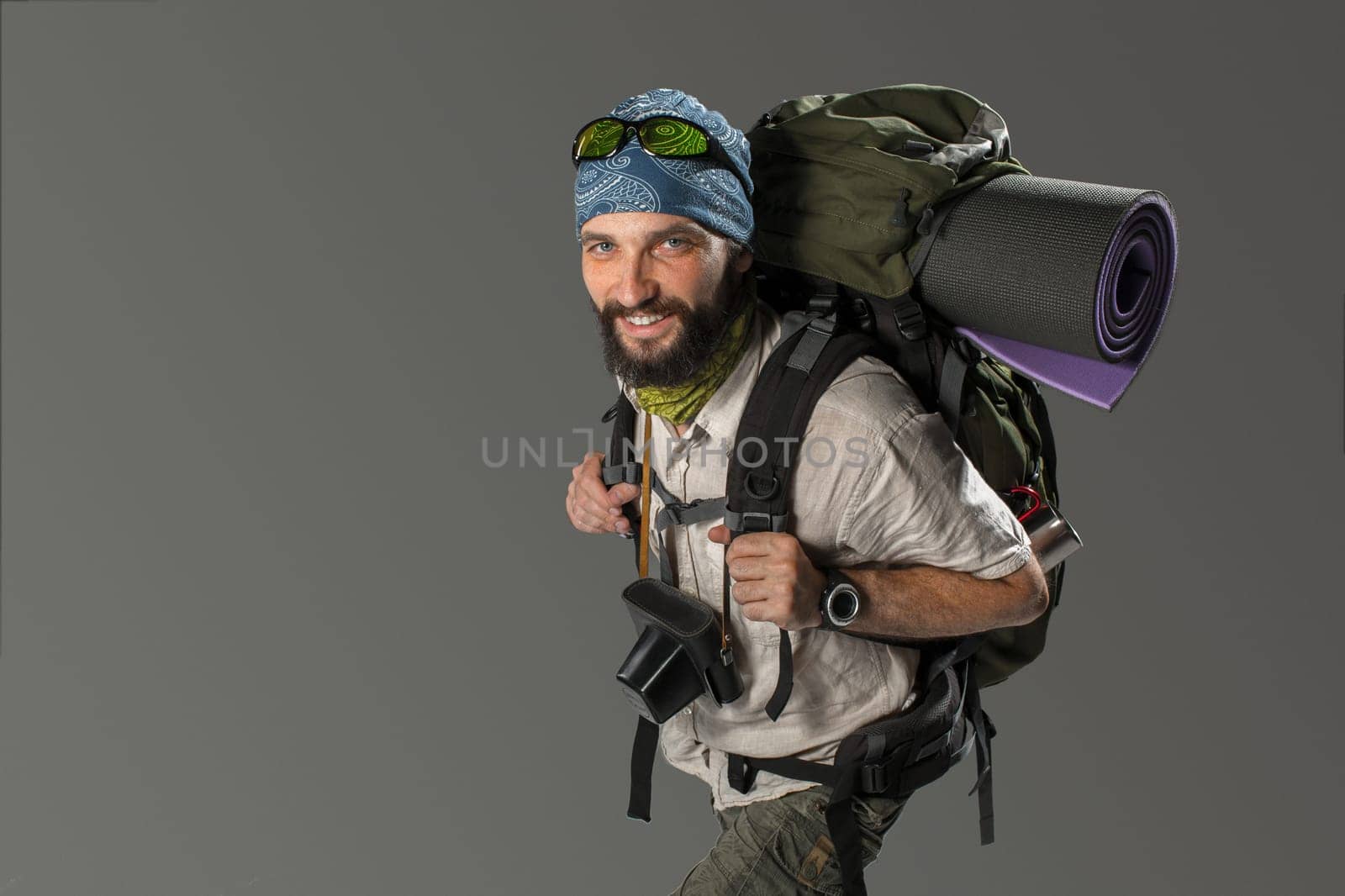 Portrait of a male smiling fully equipped tourist with backpack and the camera on gray background