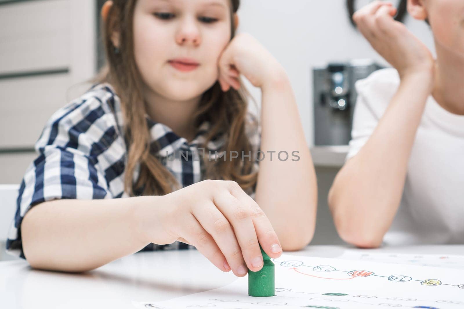 Contemplative funny little girl holding green toy paw as piece in board game above map on table board near unrecognizable boy sitting at table. Family strategic business games for kid development.