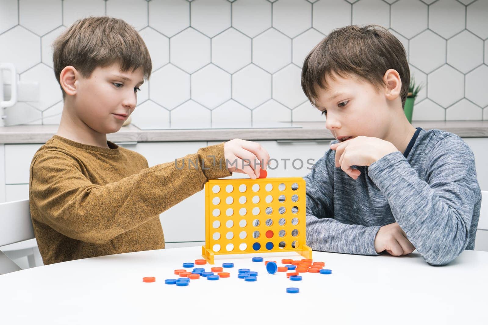 Two positive, pensive and concentrated little boys playing connect four game 3d with yellow plastic field with holes for figures on white table. Tactic and education with games. Home entertainment