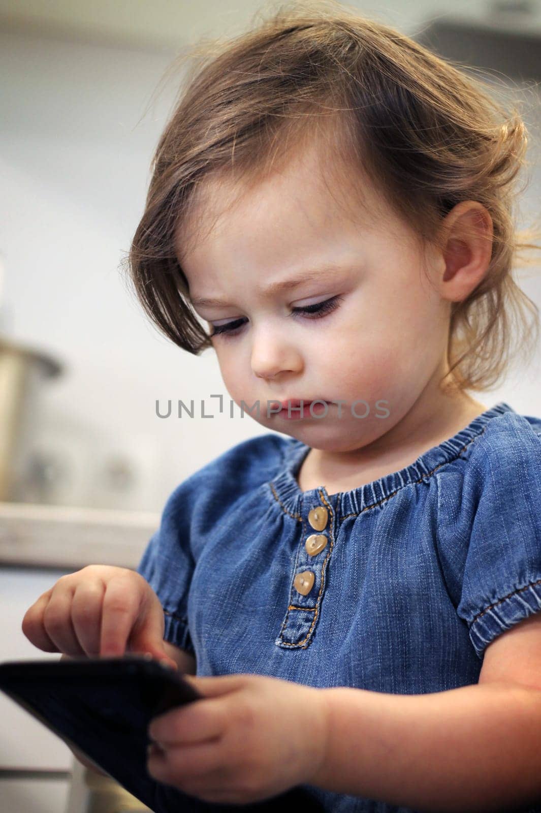 Cute little dark-haired girl 1.5-2.5 in a jeans dress types something or dials a number on a smartphone in the kitchen. Baby miss communication with relatives. Self-isolation and quarantine concept.