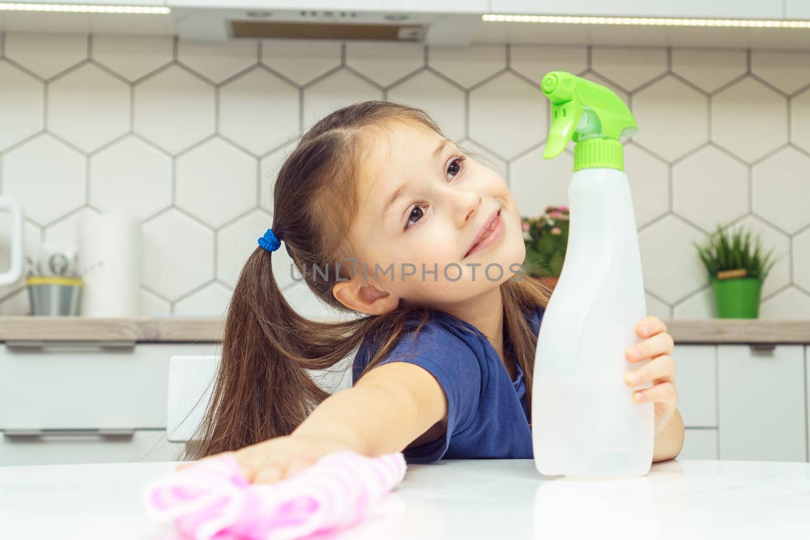 Nice little girl with detergent sprayer and household rag wipe table. Portrait of happy child tidying up kitchen, home cleaning concept. Kid help clean up house. Room cleaning supplies, cleanser.