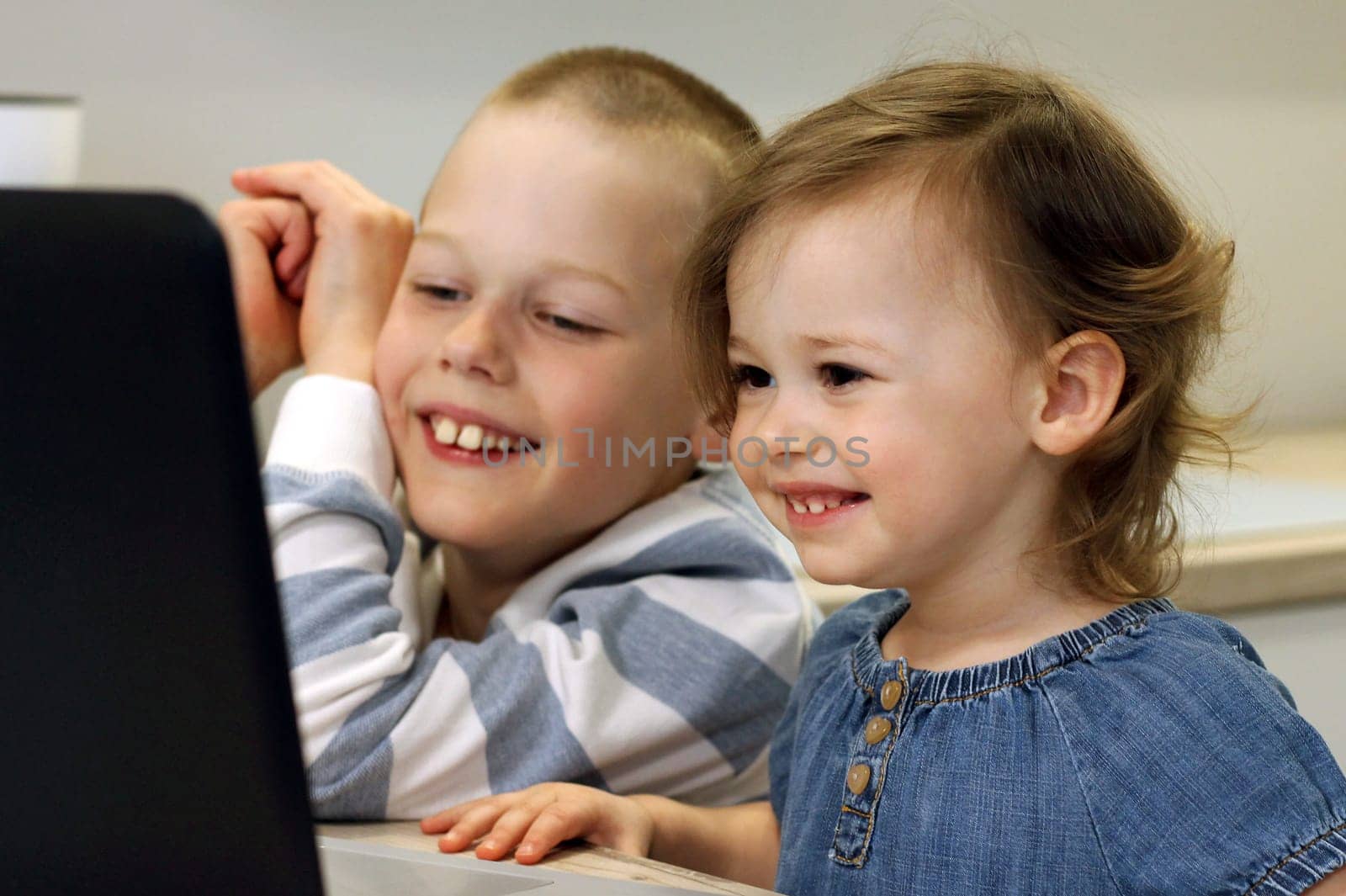 Two laughing children are sitting in the kitchen and looking at the computer. Children look at a laptop and communicate with their relatives online using the Internet. Self-isolation concept.
