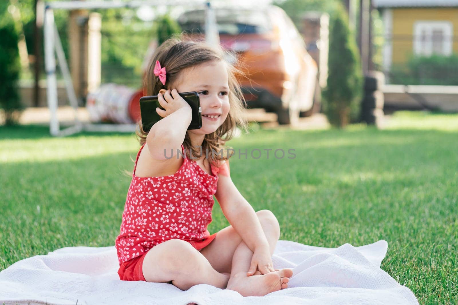 Little 3-4 girl in red clothes sits on blanket on green grass and speaks on cell phone. Children using gadgets