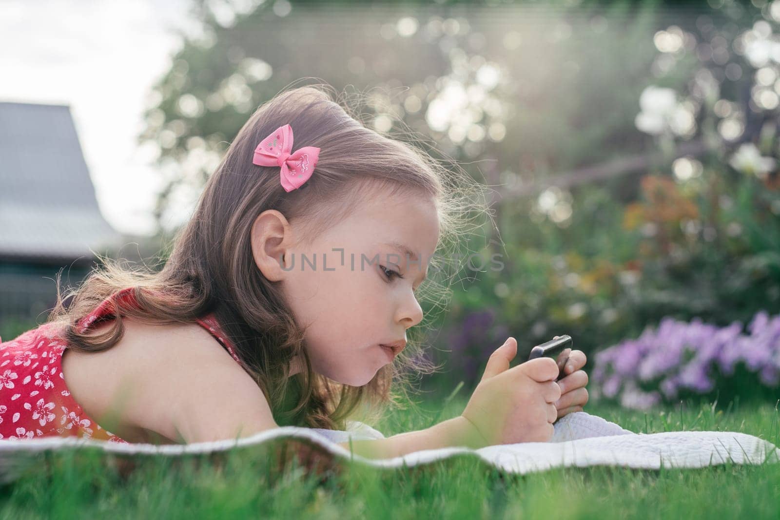 Portrait of little girl 3-4 in red clothes lying on blanket on green grass and looking into mobile phone. Children using gadgets