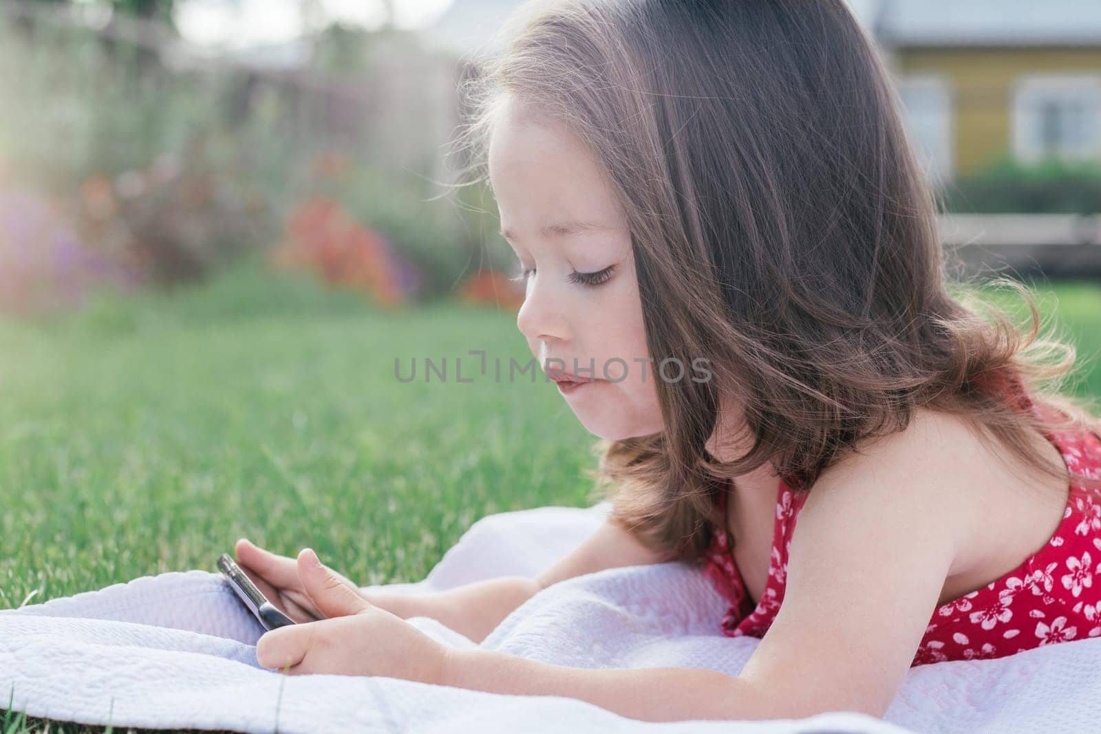 Portrait of little girl 3-4 in red clothes lying on blanket on green grass and looking into mobile phone. Children using gadgets