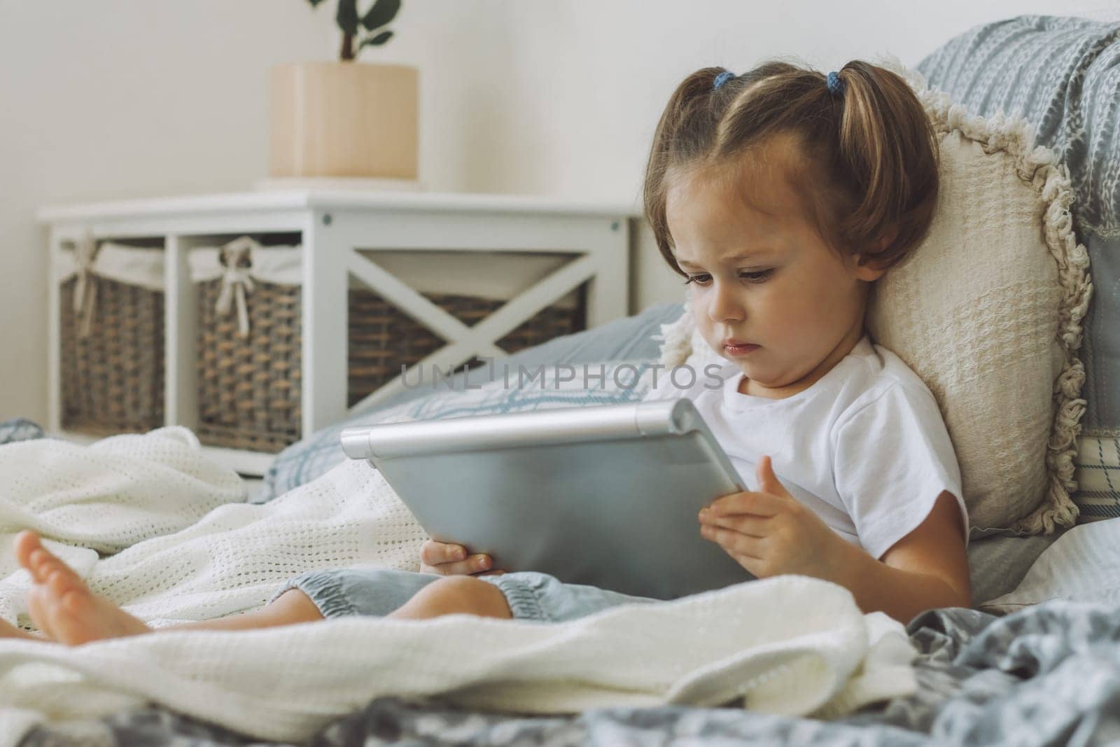 Little dark-haired girl 2-4 with two ponytails sits on bed with pillows seriously looking at tablet computer