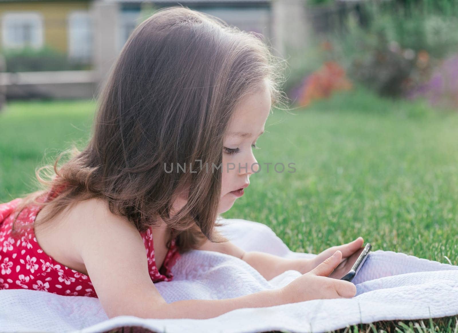 Portrait of little girl 3-4 in red clothes lying on blanket on green grass and looking into mobile phone. Children using gadgets