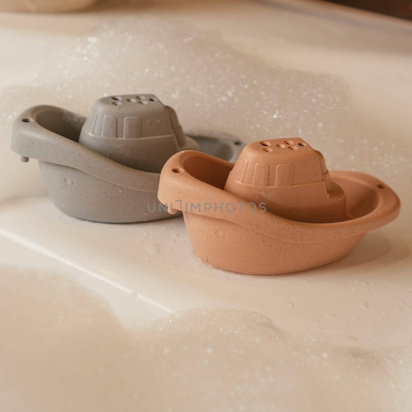 Cute grey and orange small silicone toy boats putting on edge of white bath floating among foam. Baby toys, toys for developing creativity, water games concept. Instagram use, square frame.