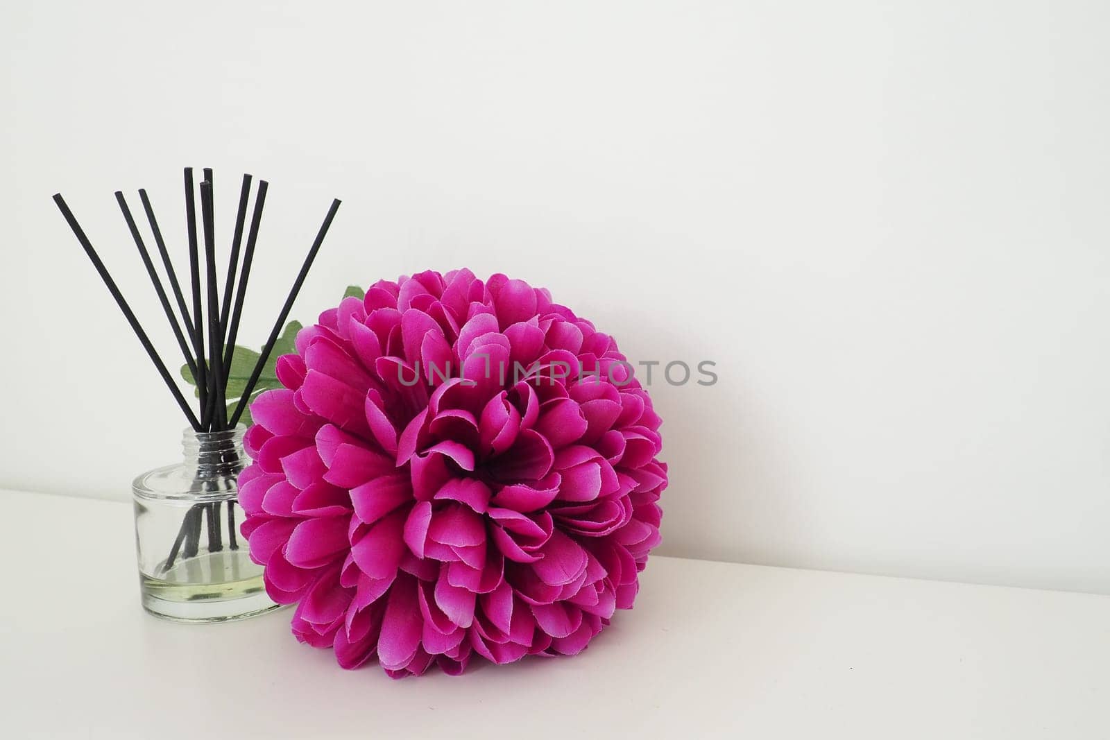 Arrangement with incense sticks, essential oil in a glass vase and a bright pink-lilac terry artificial flower in the interior of a white room. White background.