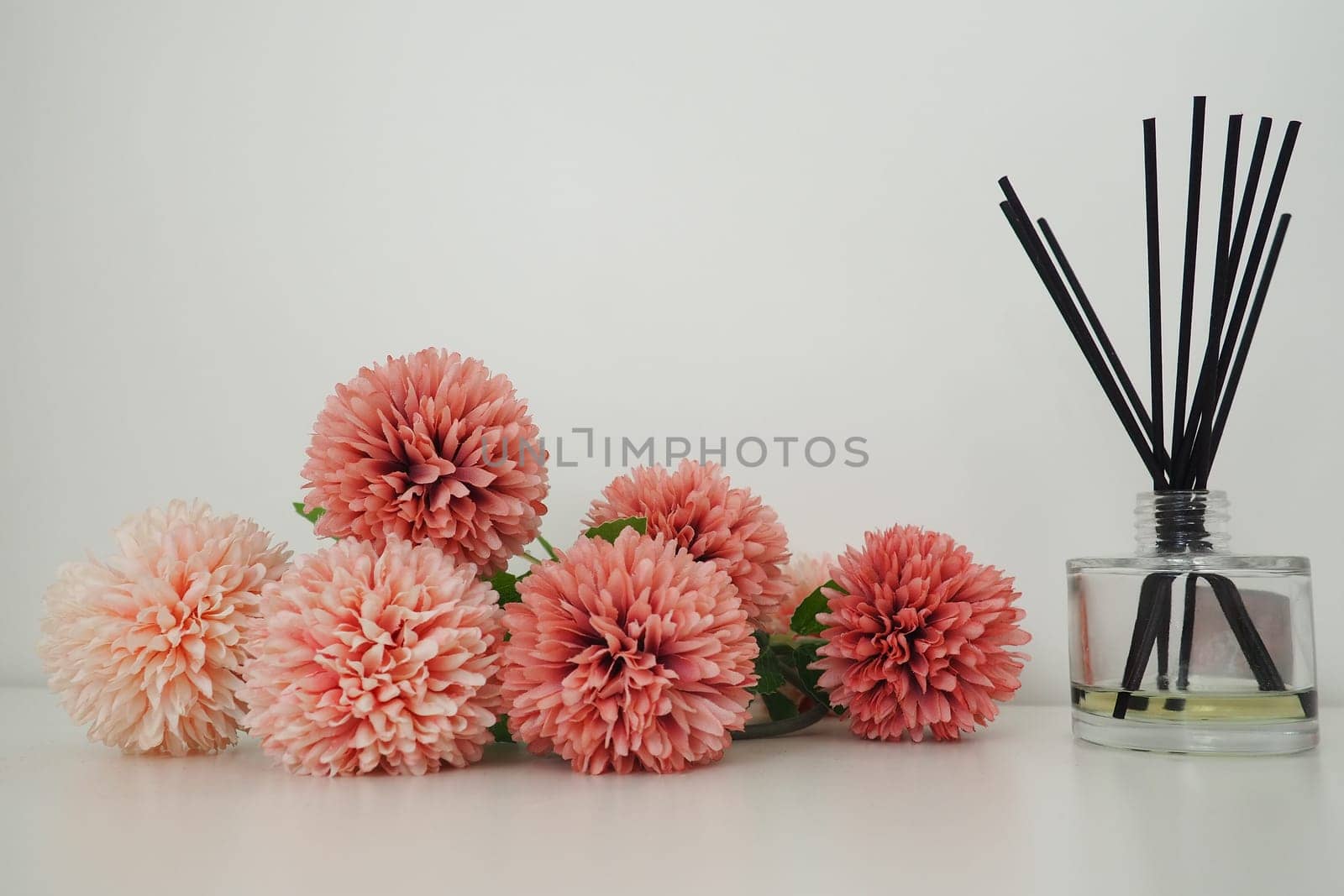 Composition with incense sticks, essential oil in a glass vase and pink terry artificial flowers in the interior of a white room. White background. Lots of flowers and scent sticks