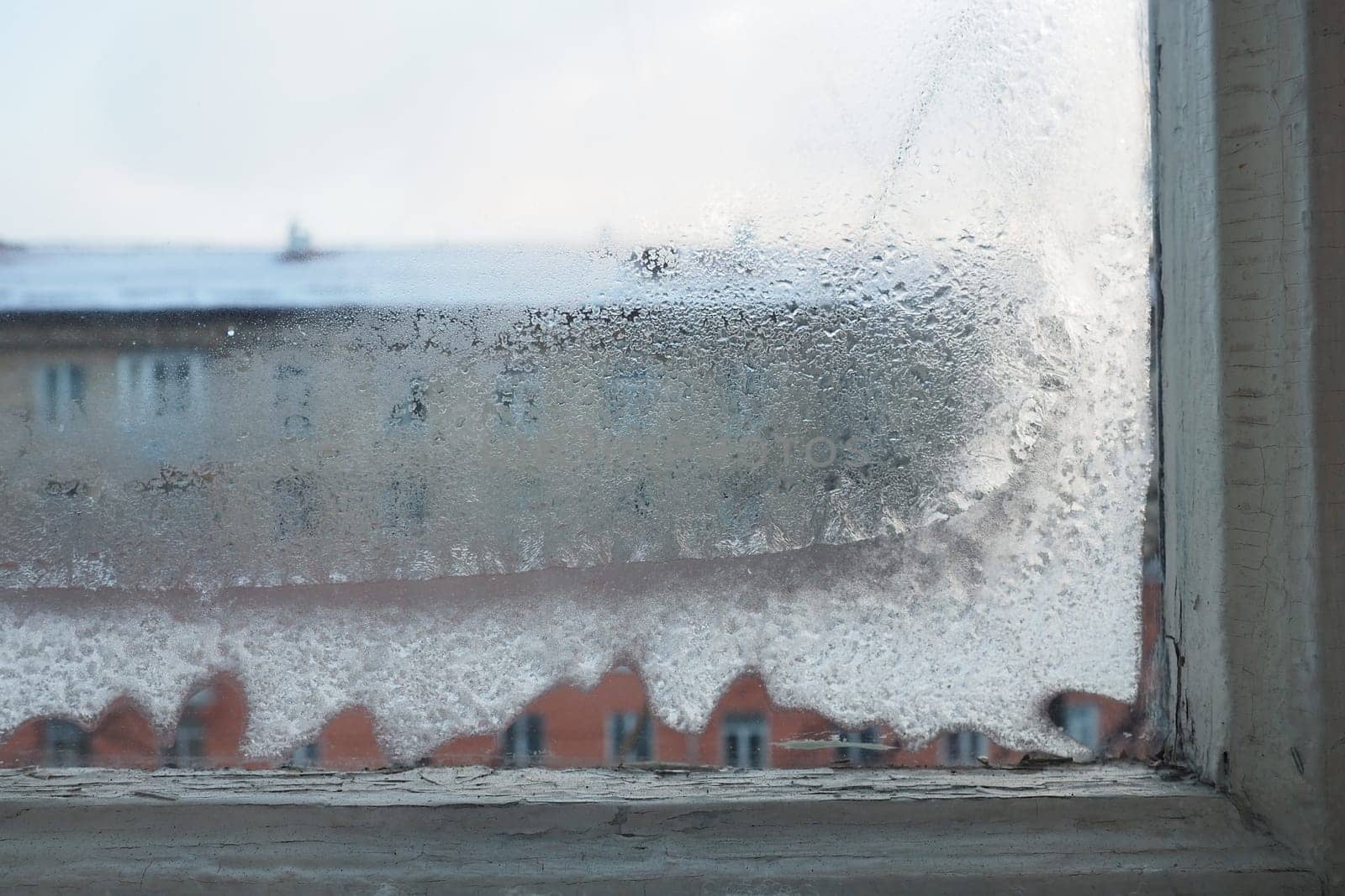 Ice, snow and frozen water crystals on glass. Window with double wooden frames. Outside the window is a street and a building. Energy efficiency and saving. Winter cold economic crisis. Freezing city.