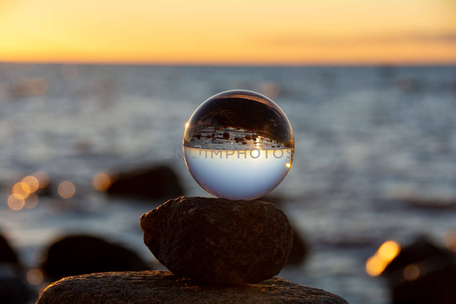 Glass ball lies on the top stone of stacked stones, at sunset on the beach, the sea and the setting sun are reflected in the ball