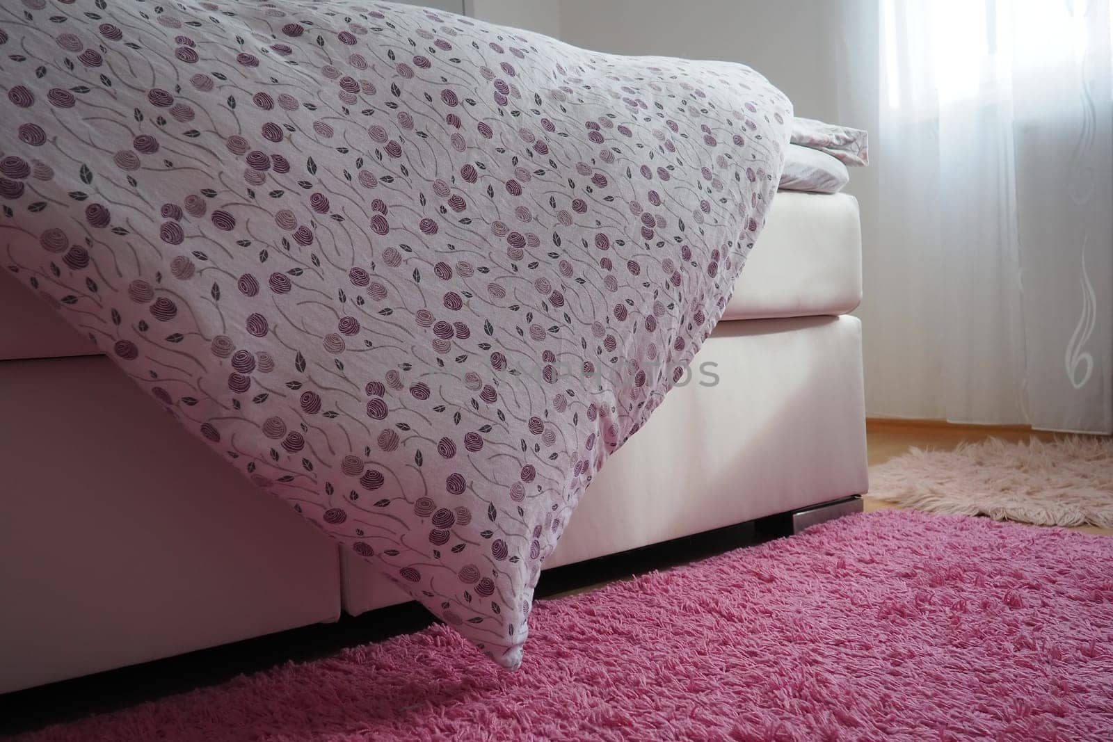 Cozy compact bedroom, white furniture, pink carpet and tulle curtains. Fluffy feather blanket thrown over the double bed. Romantic morning. Light from the window. The blanket hangs down to the floor