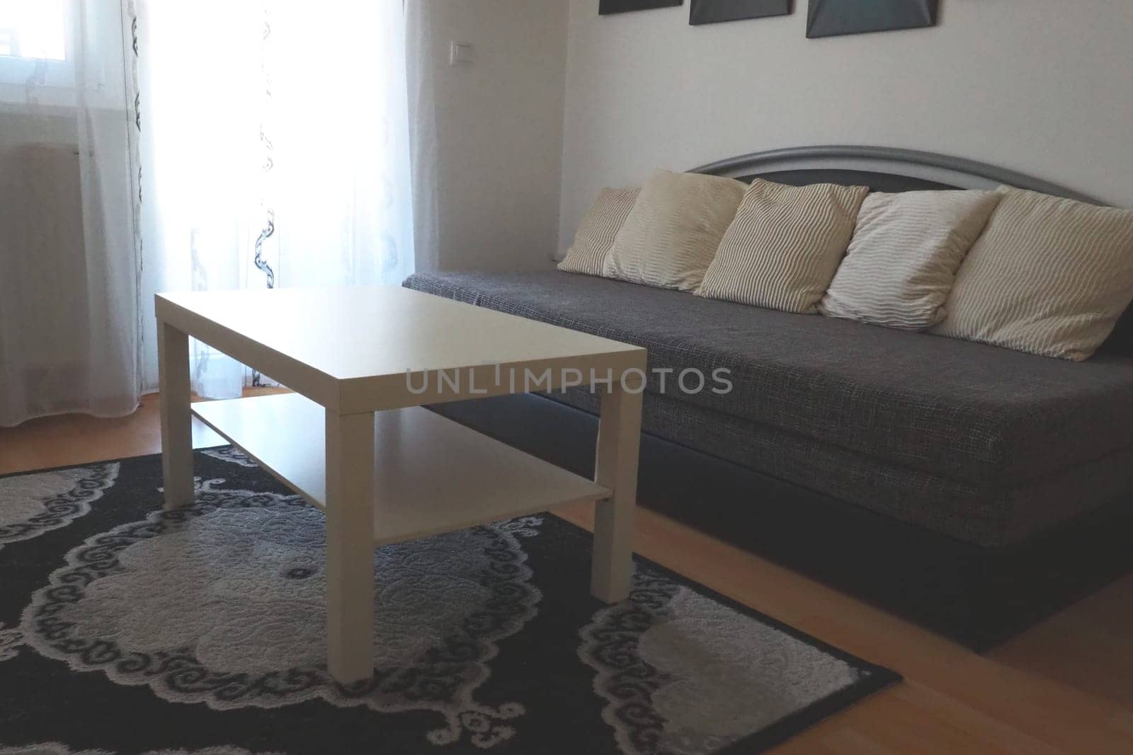 A white coffee table and a large gray sofa with decorative white cushions in the living room of a trendy apartment. Translucent tulle on the window or door from the balcony. Laminate and carpet