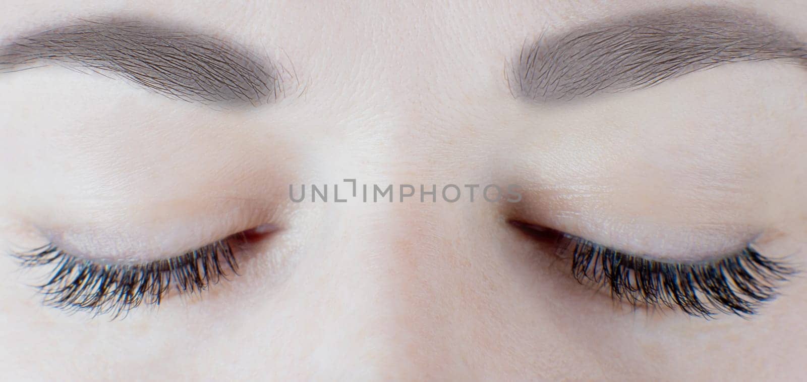 Close-up of closed eyes with long false eyelashes, black eyeliner, flawless skin, gorgeous eyebrows of unrecognizable young woman. Macro shot. Beauty procedures, cosmetology, microblading, make-up.