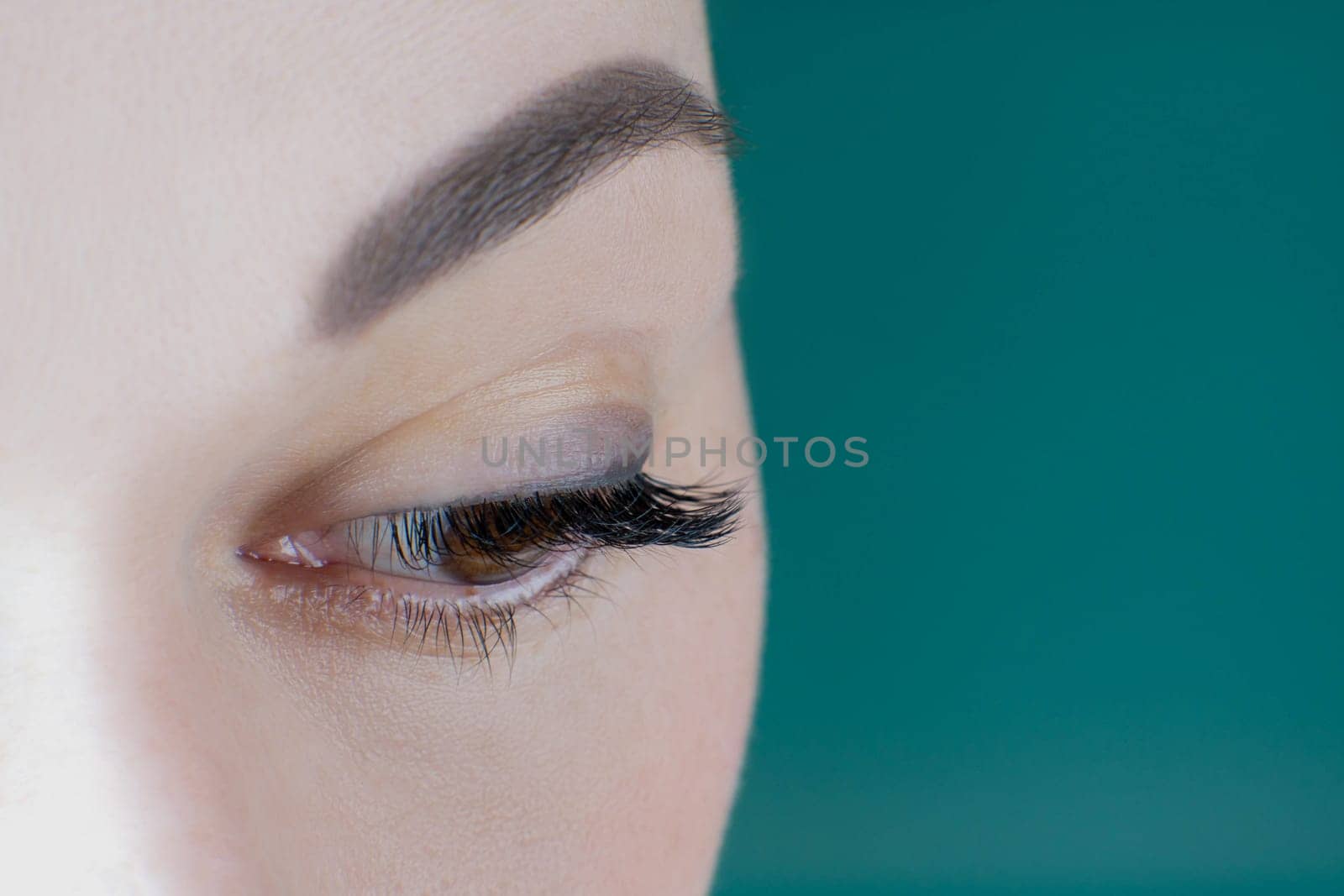 Close-up of brown eye with long eyelashes black eyeliner flawless eyebrow of unrecognizable young woman looking down on green background. Macro. Eyelash extension, cosmetology, make-up, advertisement.