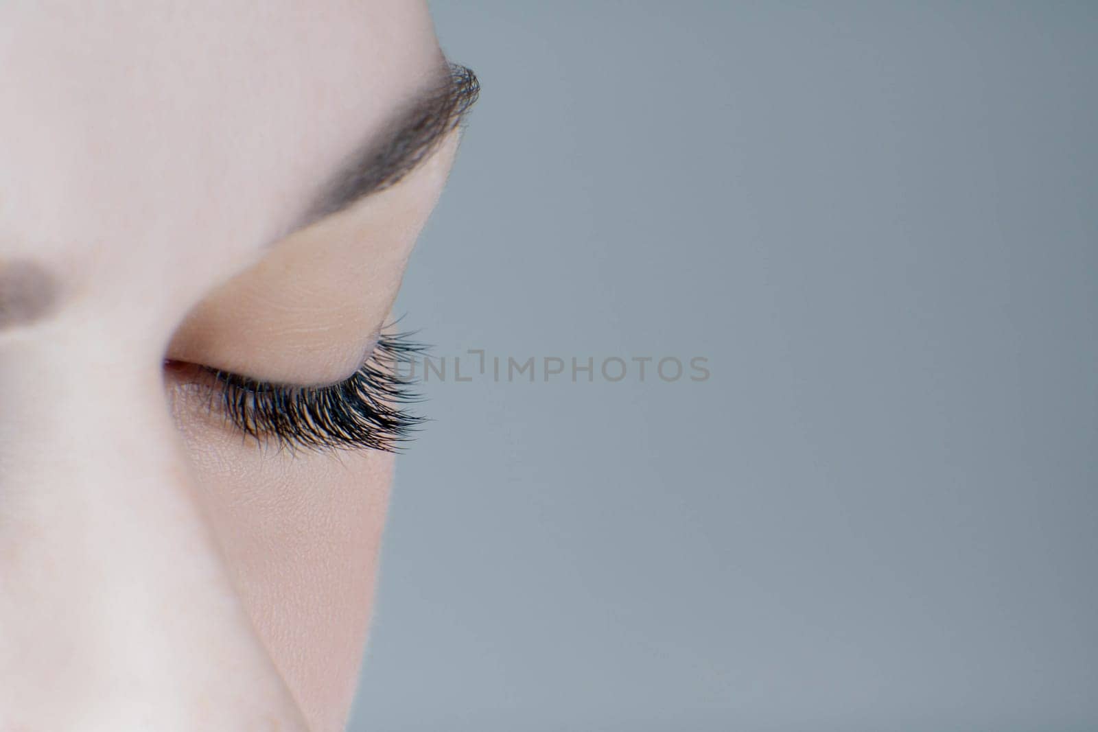 Close-up of closed eye with extreme long eyelashes, healthy skin, perfect eyebrow of unrecognizable young woman on grey background. Macro. Eyelash extension, beauty procedures, make-up, microblading.