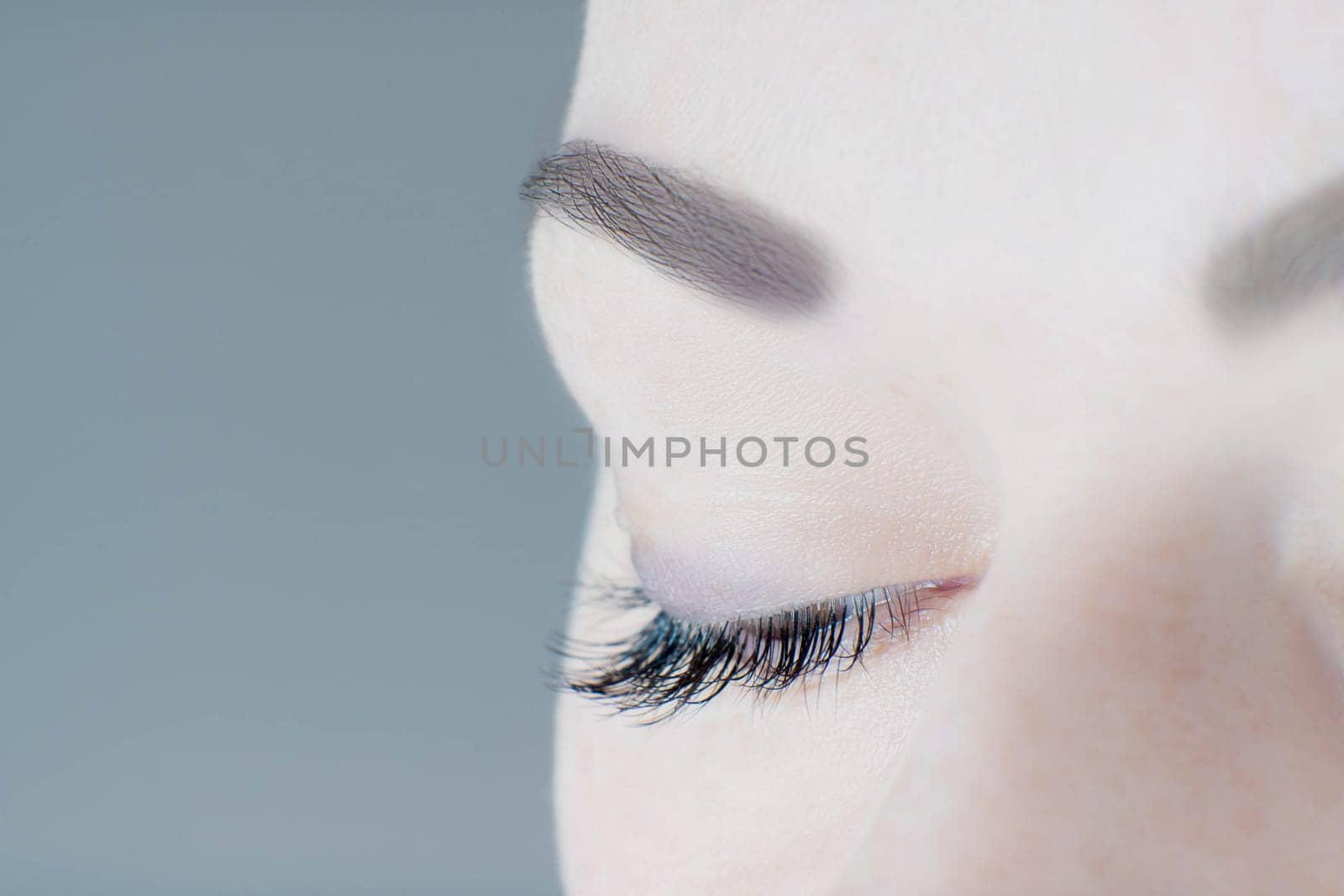 Close-up of closed eye with long artificial eyelashes, amazing skin, ideal eyebrow of unrecognizable young woman on grey background. Macro. Eyelash extension, beauty procedures, make-up, microblading.