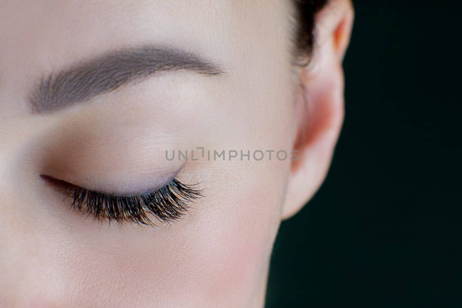 Close-up of closed eye with amazing long eyelashes, black eyeliner, perfect skin, eyebrow of unrecognizable young woman on black background. Macro. Eyelash extension, beauty procedures. Soft focus.