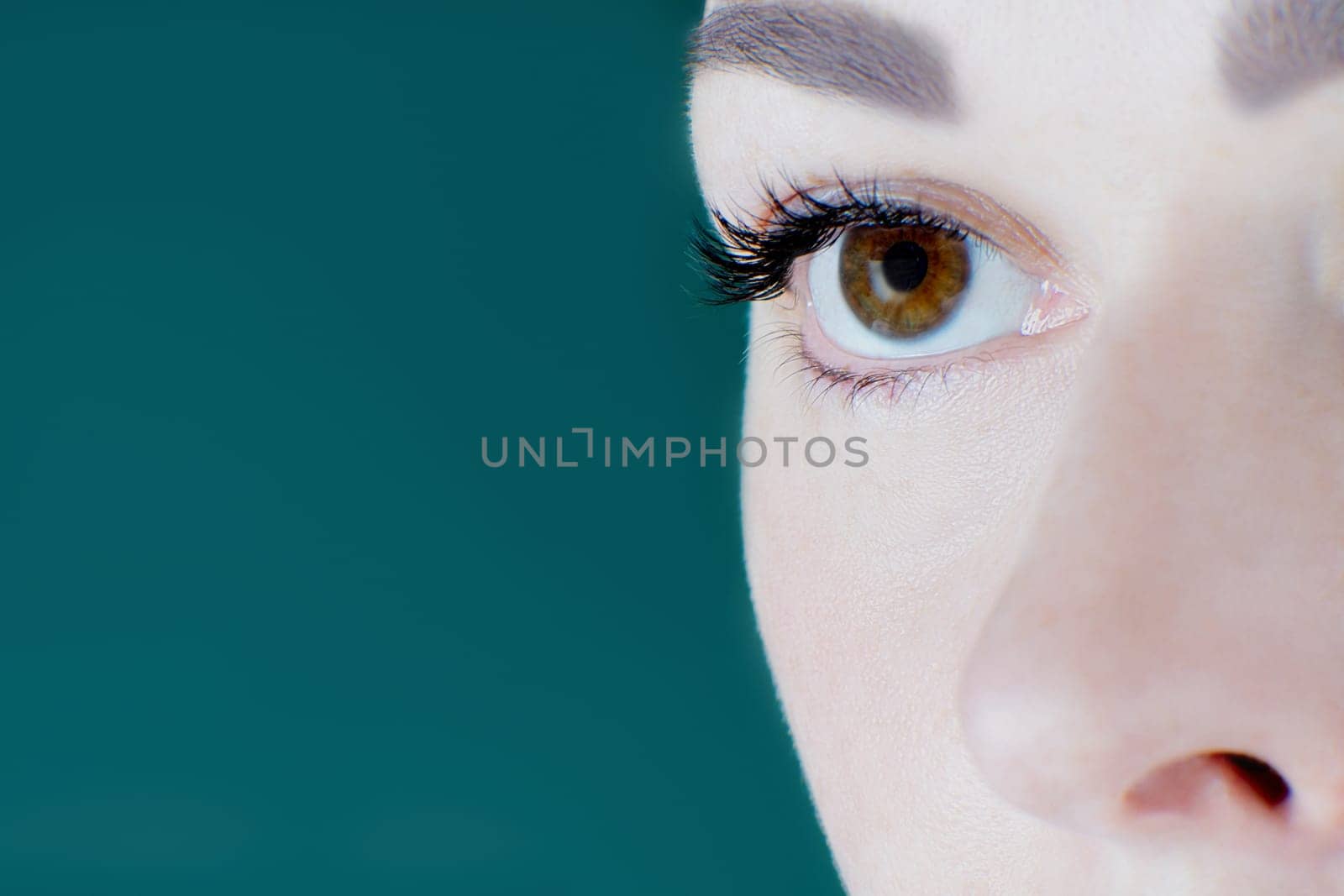 Close-up of brown eye with artificial eyelashes, healthy skin, eyebrow of unrecognizable young woman looking aside on green background. Macro. Eyelash extension, cosmetology, make-up, advertisement.