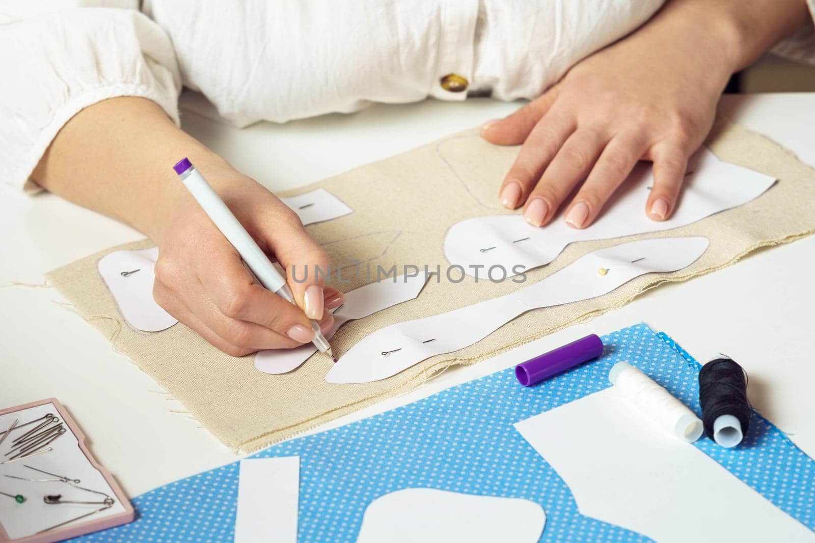 Cropped female seamstress drawing contours with pen of pinned paper sewing patterns on textile sheet on table. Needlework, making patterns for future items such as toys or clothes. Small business.